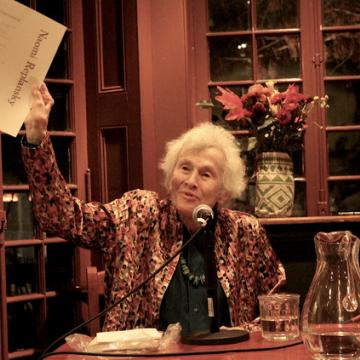 Naomi Replansky at the Kelly Writers House, speaking into microphone while holding a paper in the air for audience to see